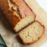 Close-up of a slice of banana bread with two ripe bananas beside it on a wooden table.
