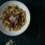 Overhead view of a bowl of pasta with meat sauce and grated cheese on a dark wooden surface.