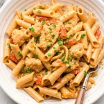 Overhead view of creamy penne pasta with vegetables and parsley in a white speckled bowl with a serving spoon.